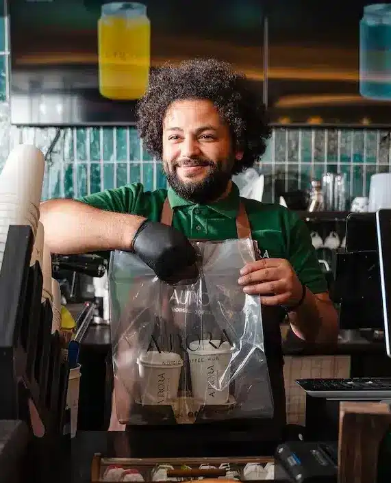 Barista at Aurora Coffee Hub handing over a takeout bag with two coffee cups.