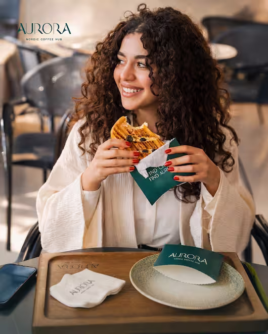 A smiling woman enjoying a freshly grilled, toasted pan-pan sandwich in branded green packaging at an outdoor table of Aurora Coffee Hub.