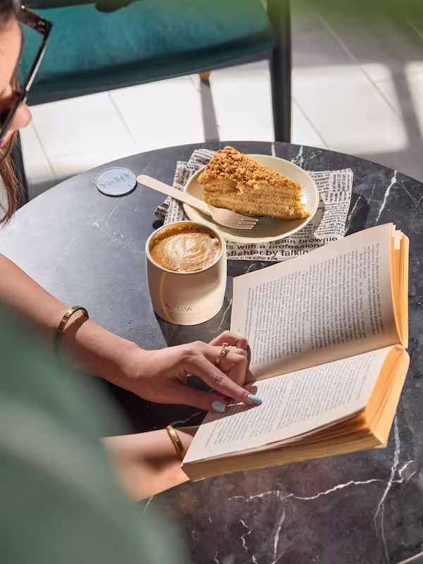 A customer reading an open book at a black marble table with a hot latte in an Aurora cup and a slice of homemade Scandinavian cake at Aurora Coffee Hub.