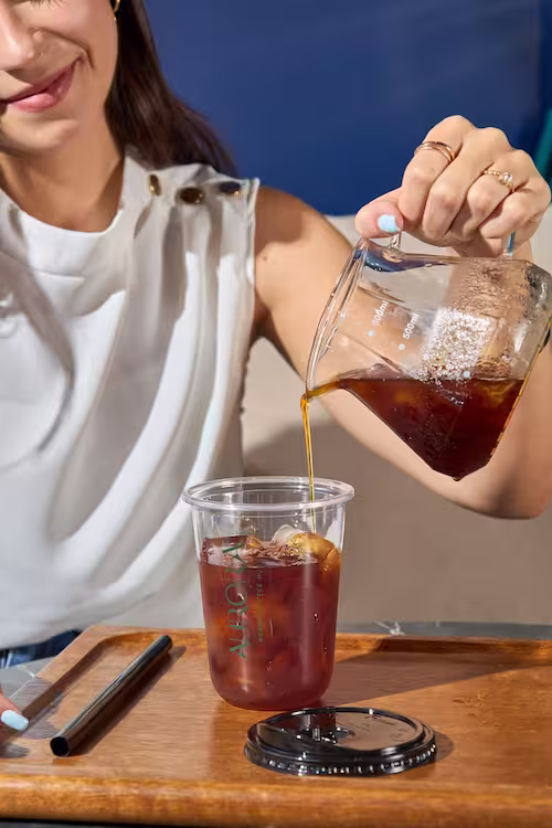 A woman pouring freshly brewed specialty coffee from a glass server carafe into a clear cup filled with ice on a wooden tray.