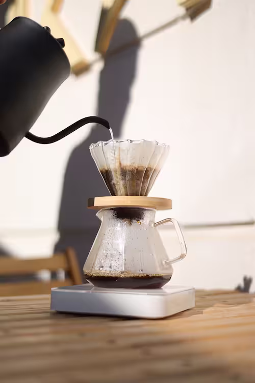 Hot water pouring from a matte black gooseneck kettle into a glass V60 dripper set over a coffee server on a digital scale in outdoor sunlight.